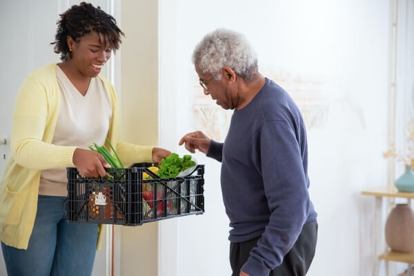 Woman holding a crate of vegetables for elderly man to pick from