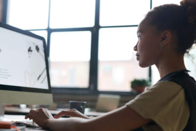 Woman working at a desktop computer in an office, focused on the screen with a large window in the background.